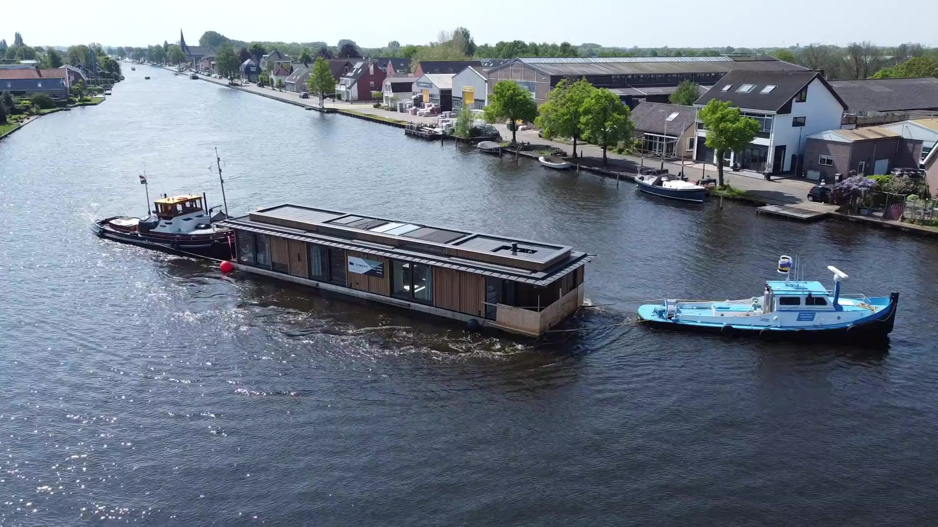 Luchtfoto van een sleepboot die een nieuwe drijvende woning met Domera-doek over het water trekt, met een tweede sleepboot erachter