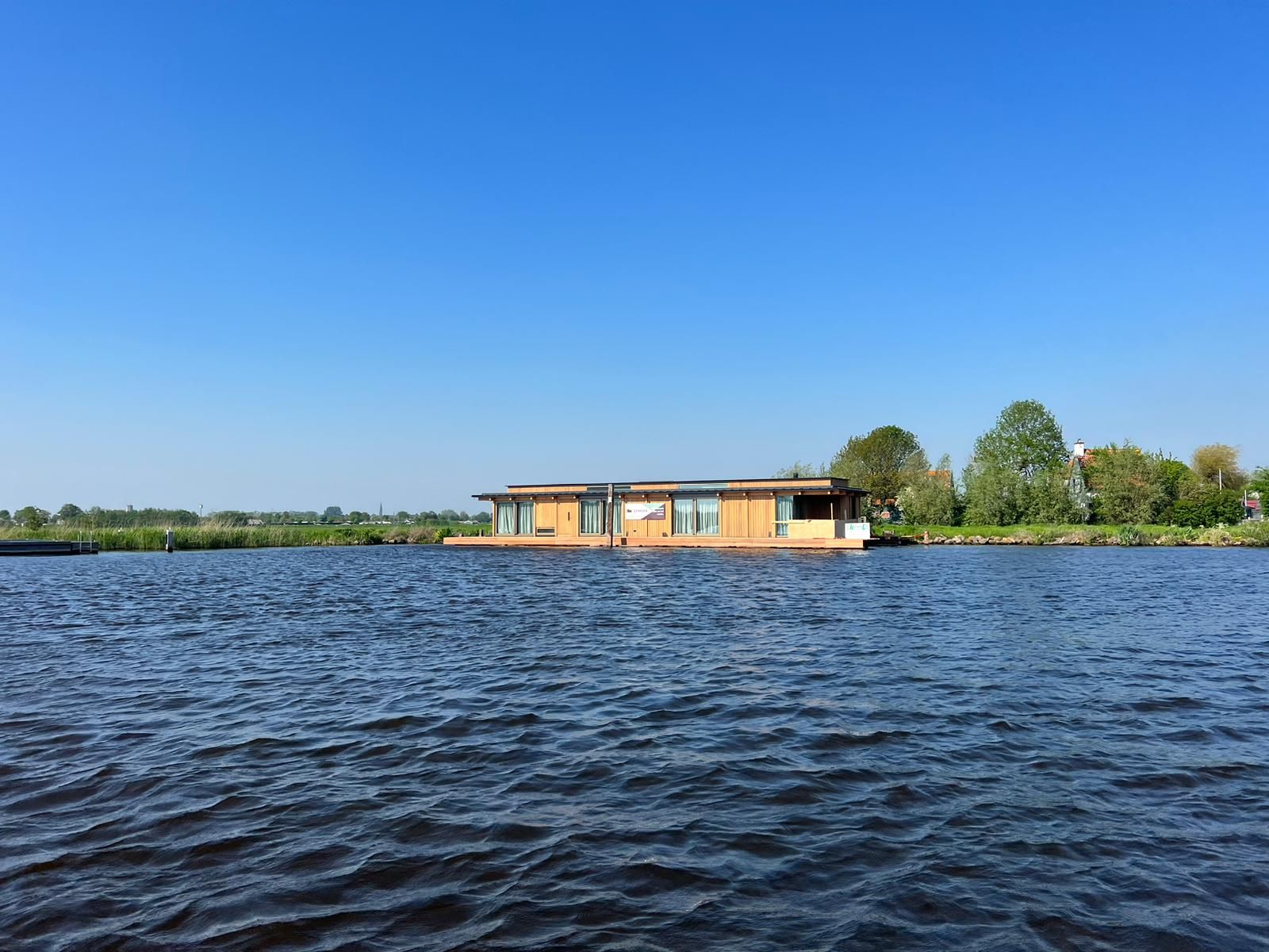 Drijvende woning aan een polderligplaats onder een blauwe lucht, gezien vanaf het water