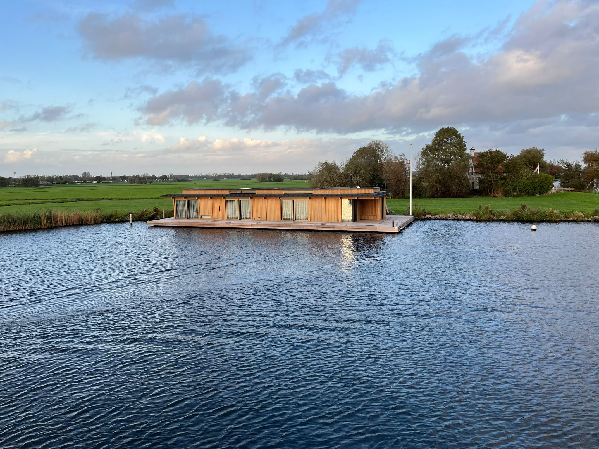 Drijvende woning met een houten gevel aan een polderligplaats in het avondlicht, met breed open water ervoor