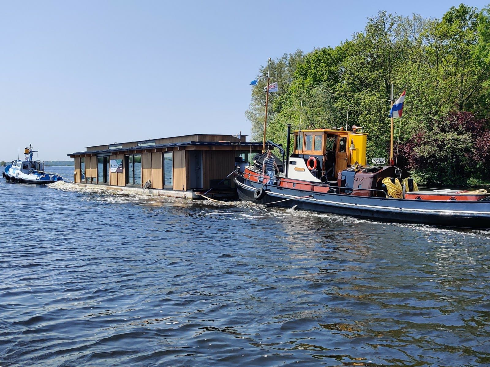 Sleepboot met bemanning trekt een nieuwe drijvende woning langs een bomenrijke oever