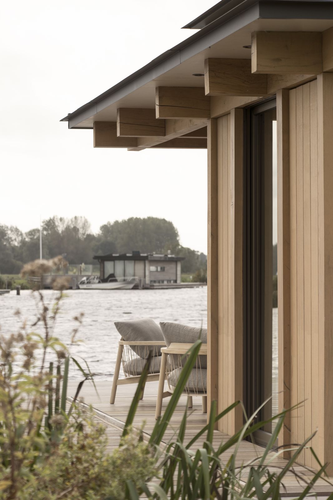 Twee tuinstoelen op het terras onder de overstek, met riet en water op de voorgrond