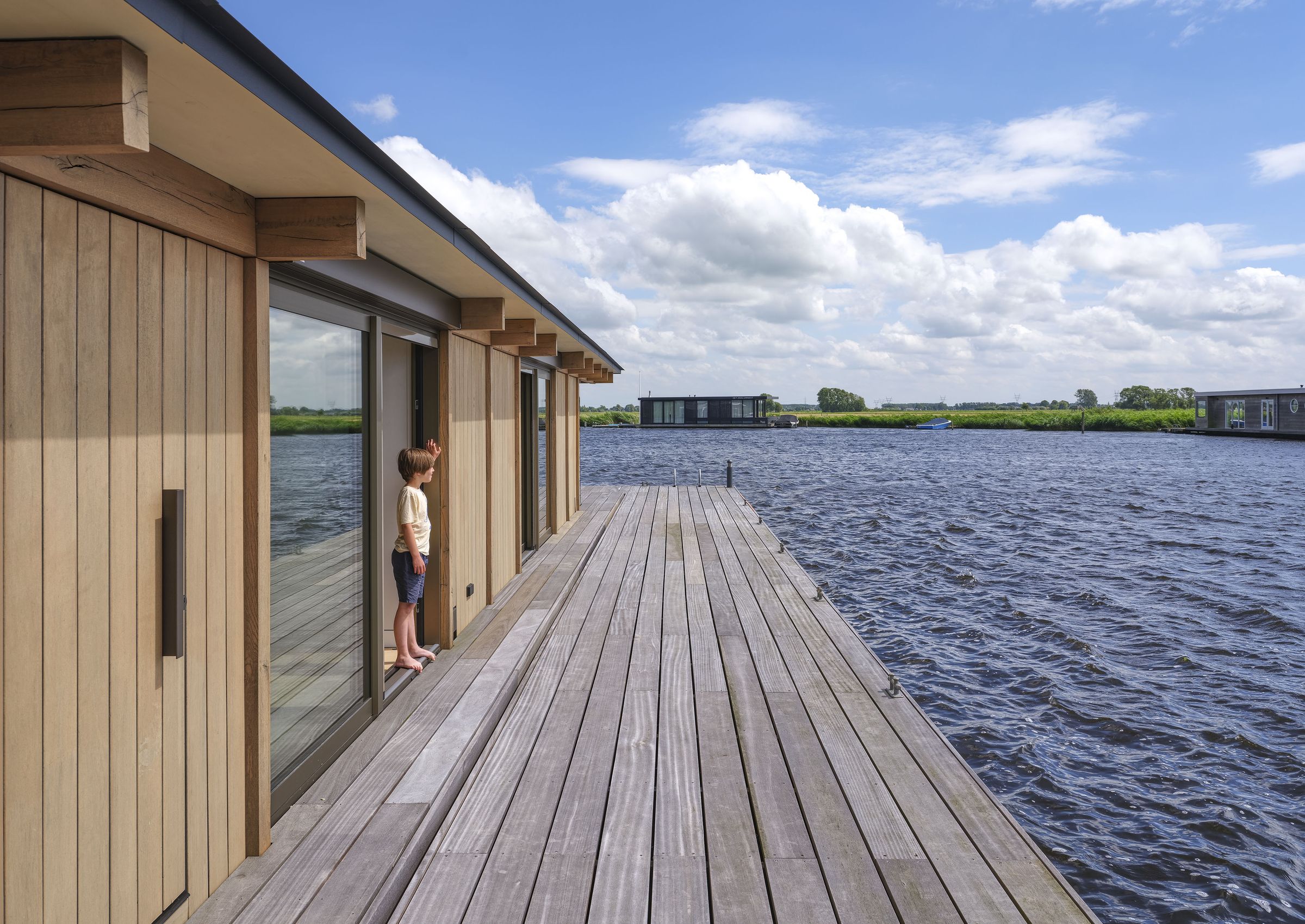 Breed houten loopdek langs een glazen gevel, met uitzicht over het water